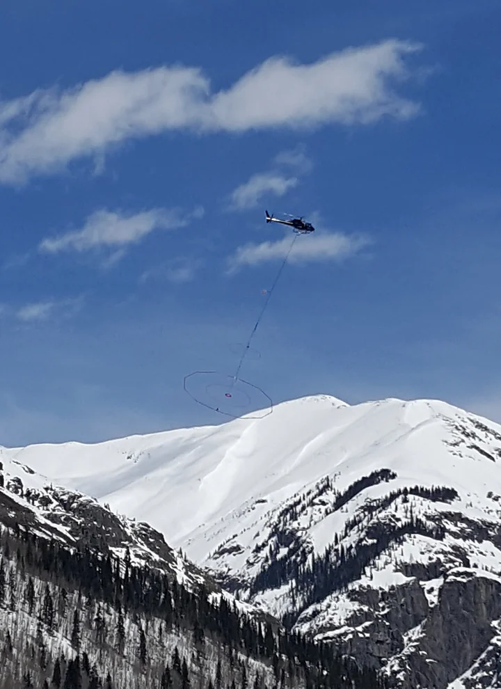 A civilian helicopter carrying geophysical survey equipment above snowy mountains in Colorado.